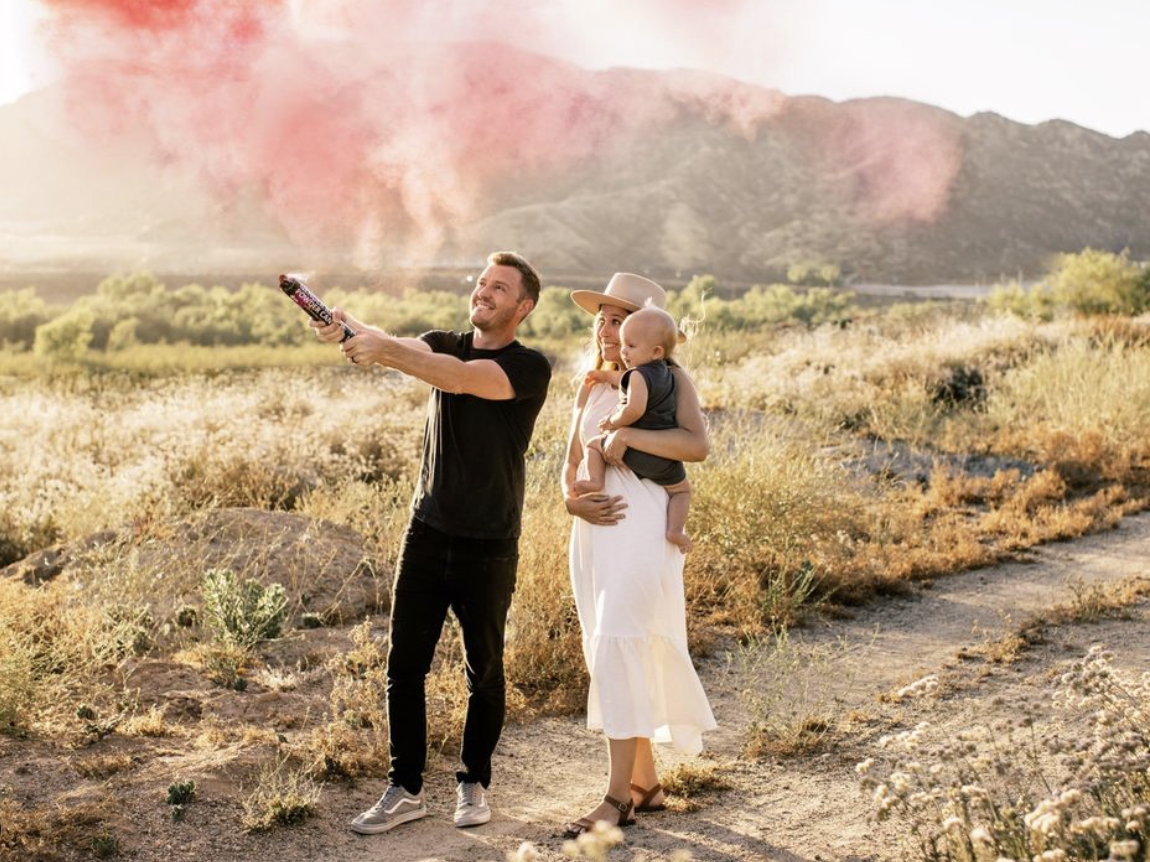 a heterosexual couple and baby popping a colour cannon to reveal the 'gender' of their next child. occuring in a desert and the canon reveals pink.