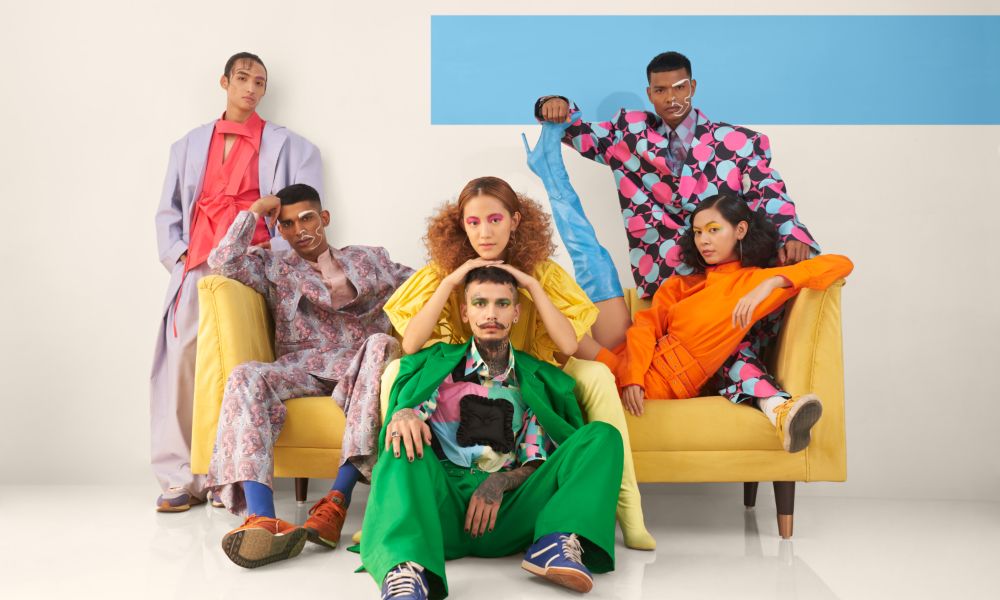 model photo shoot, six poeple three sitting on a yellow couch and two standing behind couch, one holding the person on the right of the couches leg. one sitting in front of the couch. editorial models shoot. 