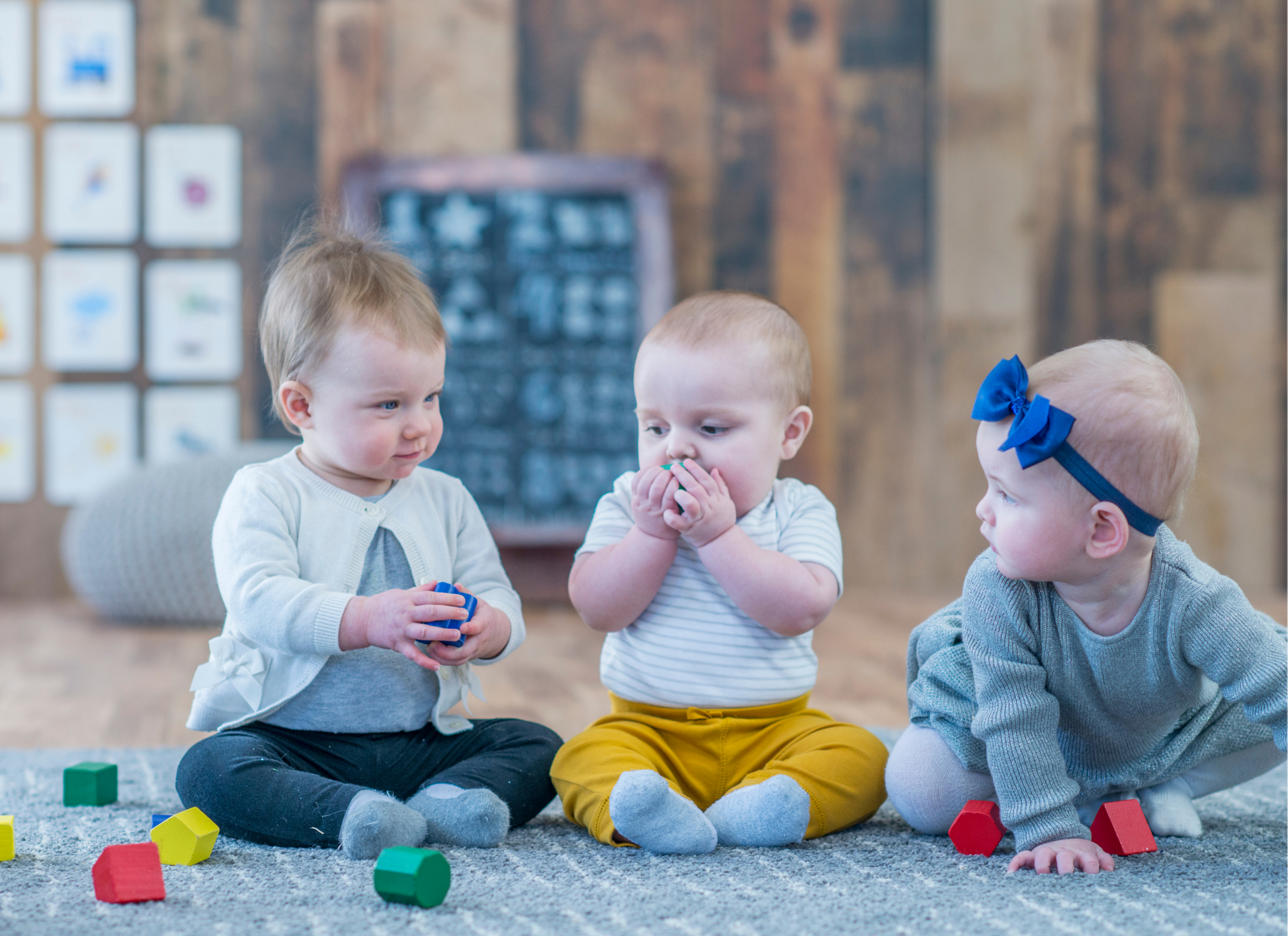 image of three 1 year olds sitting up on a blue rug. background is a kindy with wooden walls and drawings on the wall. the babies are playing with bricks