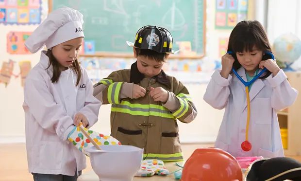 three children playing dress up in a classroom. the girl on the left is dressed up in a chefs hat and coat. she is playing with a mixing bowl and spoon while putting on a rainbow oven mit. the middle boy is putting on a firefighters outfit and wearing a hardhat. the girl on the right is playing with a stethoscope and dressed in lab coat.