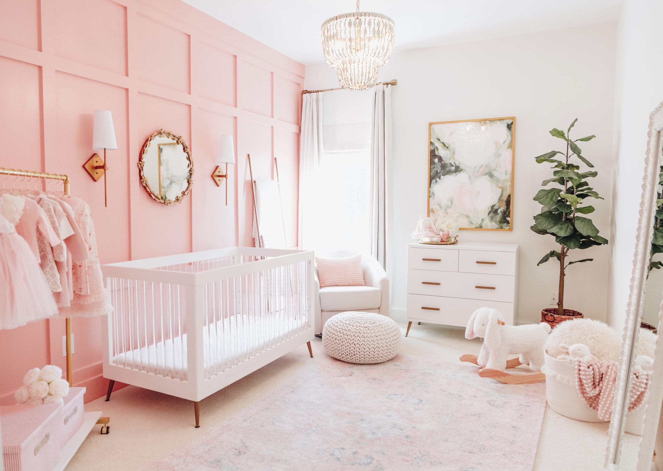 stereotypical young girls room. walls painted pink, with chandallier, dresses handing in left foreground, next to it a pink cot, then a pale pink armchair and fouton. white set of draws, bunny rocking chair and mirror in right foreground.