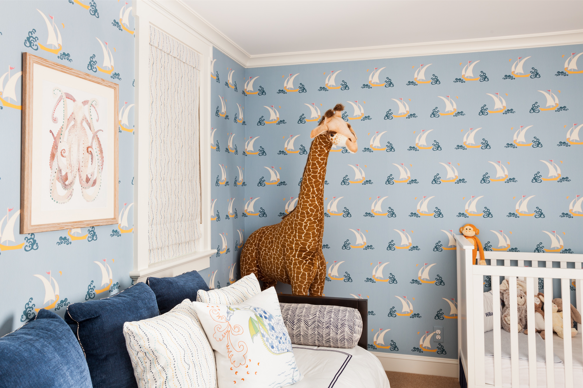 stereotypic young boys room. blue wallpaper with sailboats, a giant giraffe in middle of scrren (background) next to it on the right is a white cot. in the foreground is a blue dressed daybed.