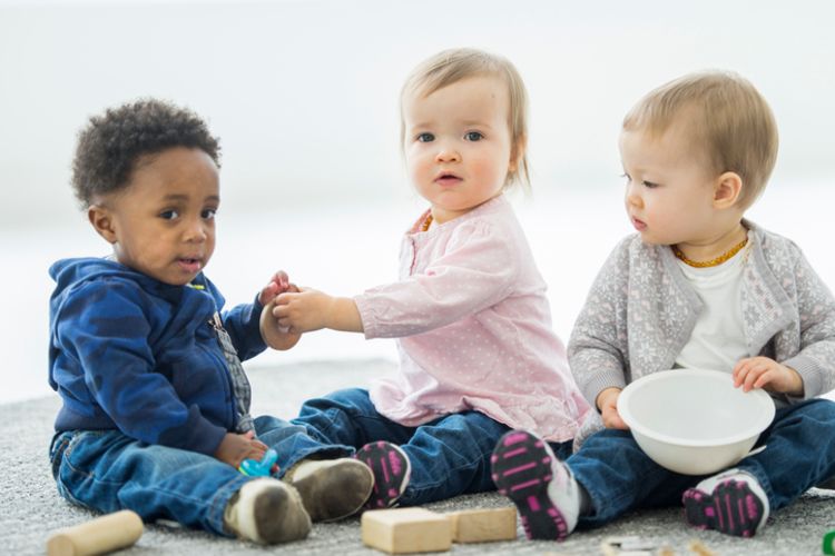 slighly older children sitting up on the floor playing with blocks, a bowl and each others hands.