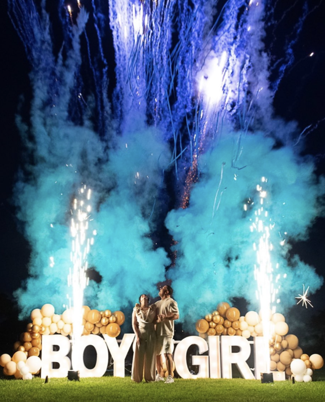 large scale gender reveal, firewords and flares in the background (blue). foreground is heterosexual couple with bif light up white boy/girl signs and gold balloons.